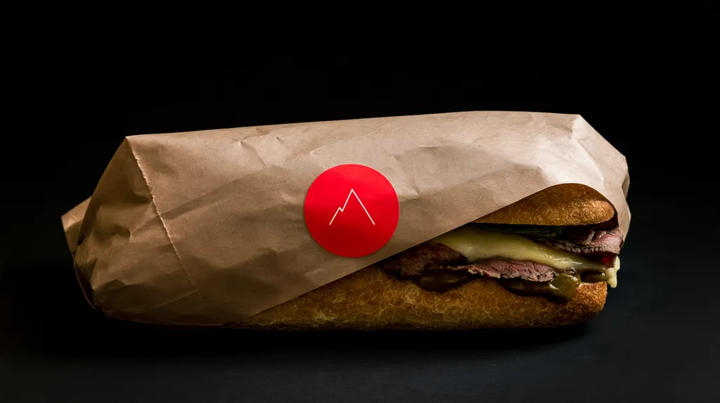 A roast beef sub half wrapped in kraft paper with a red circular mountain seal, on a black surface.