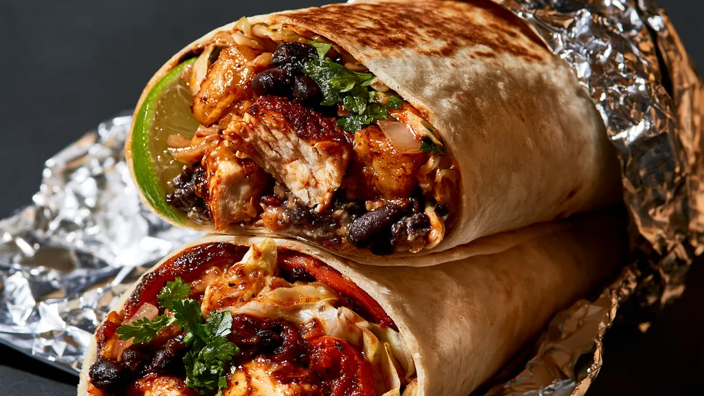 Two halves of a chile chicken burrito with black beans, shredded cabbage, cilantro and lime, stacked on crumpled foil on a black studio background.