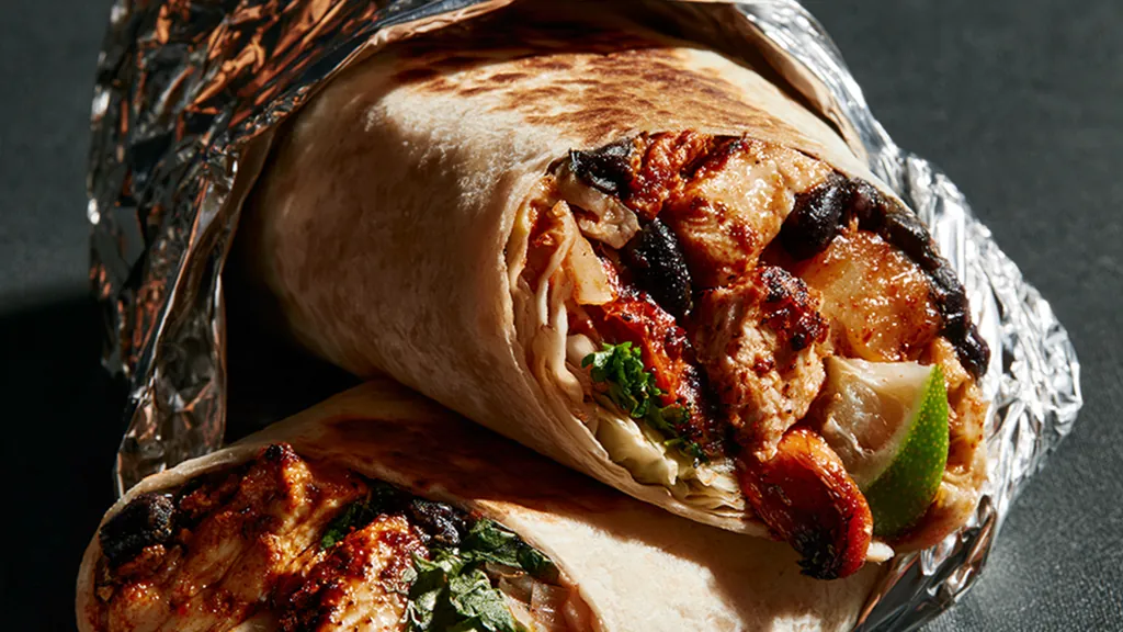 A grill-marked flour tortilla burrito cut in half and wrapped in foil, showing chile-marinated chicken, black beans, cabbage, cilantro and a wedge of lime.