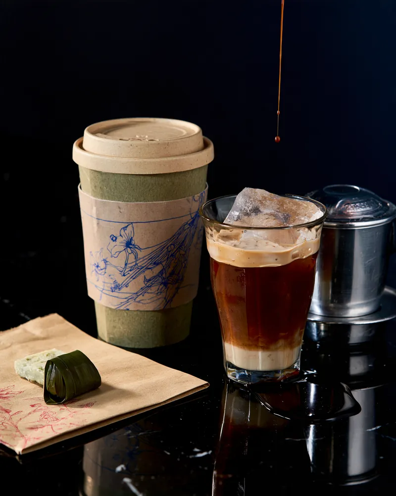 Vietnamese iced coffee layered over condensed milk beside a steel phin filter and a small pastry.